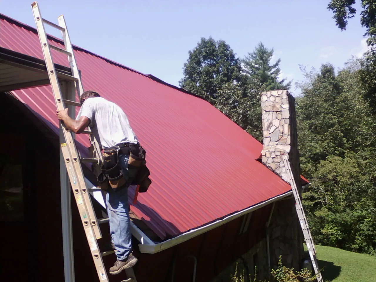 Roofer on a ladder beside a red metal roof and stone chimney — Soffit & Fascia Repair by Roofing Brothers in Oley